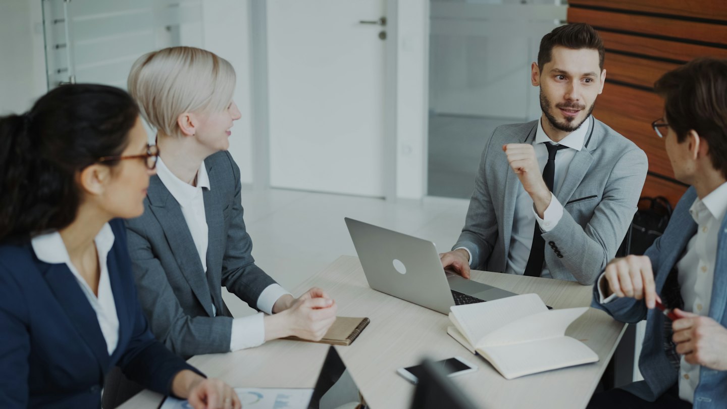 Professionals bespreken zaken aan een vergadertafel met laptops en documenten.
